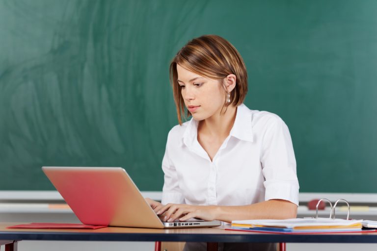 Professora sentada em sua mesa, em frente à lousa, trabalha usando seu notebook.