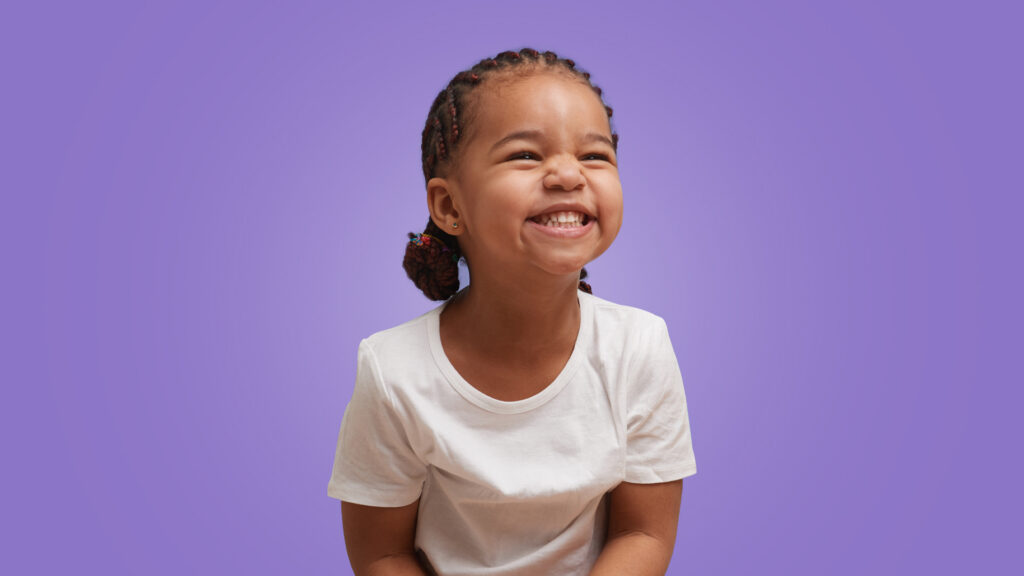 menina de 2 anos sorrindo como se olhasse para alguém. Ela tem a pele negra, cabelo trançado e veste camiseta branca em um fundo roxo.