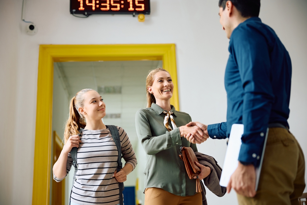 No corredor de uma escola, um diretor escolar cumprimenta com um aperto de mão a mãe de uma adolescente, que está ao lado. Todos sorriem.