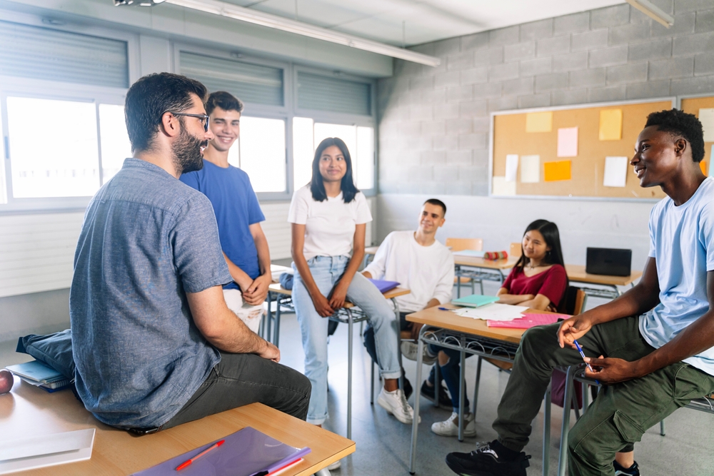 Professor sentado em cima da mesa conversa com alunos adolescentes de maneira informal em sala de aula.