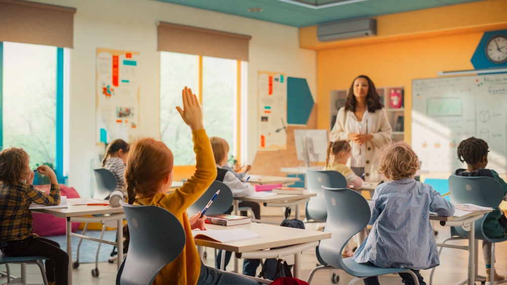 Aluna levanta a mão sentada na cadeira e professora presta atenção em frente à sala de aula.
