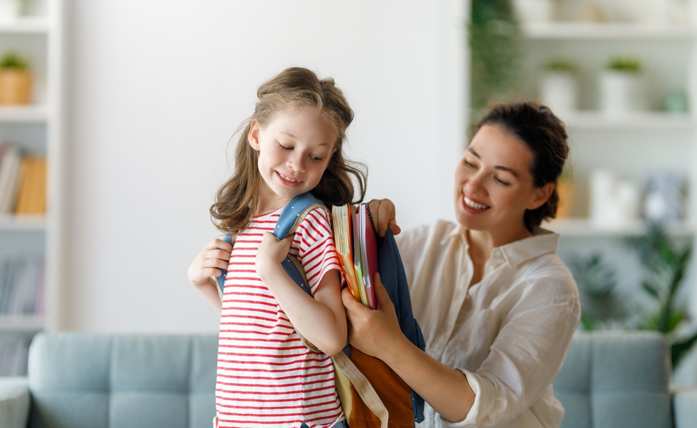 mãe ajuda filha pequena a colocar os materiais na mochila para ir à escola.