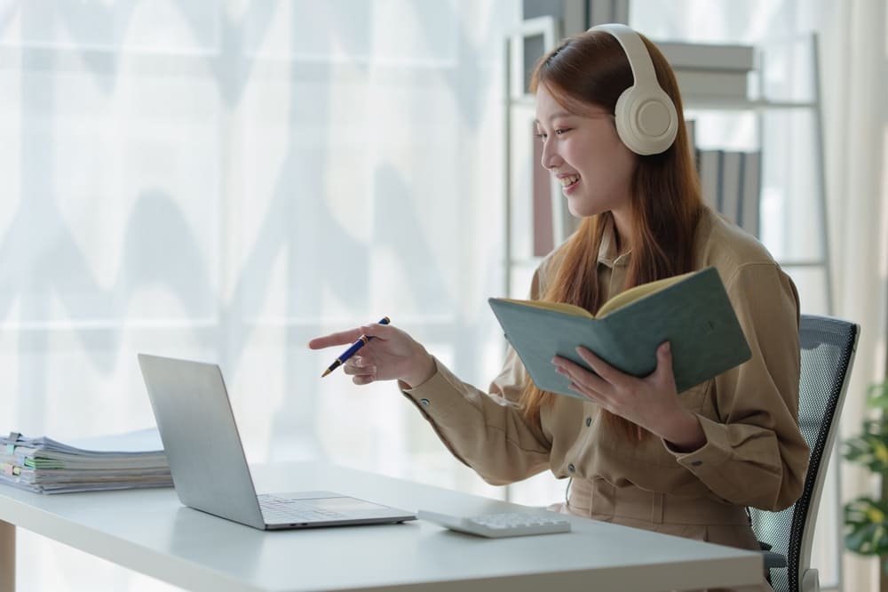 Mulher com fones de ouvido estuda inglês online com caderno e notebook sobre a mesa.