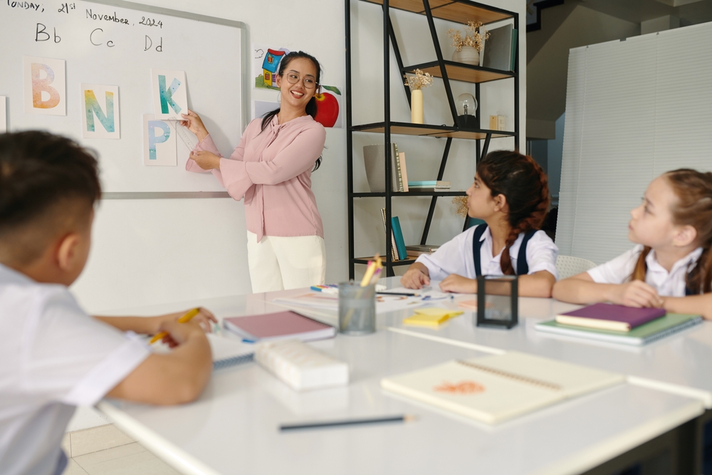 Professora apontando letras do alfabeto em um quadro enquanto crianças assistem atentas.