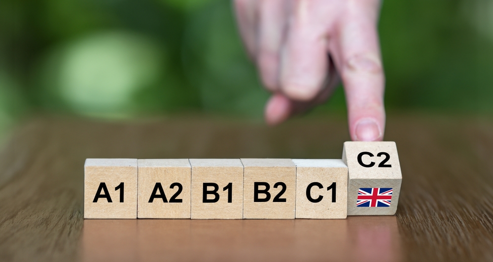 Blocos de madeira com os níveis de inglês A1, A2, B1, B2, C1 e C2, com uma mão apontando para o bloco C2.