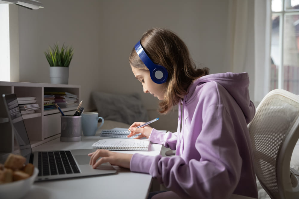 Estudante assistindo a uma aula remota no laptop, anotando em um caderno, usando fones de ouvido.