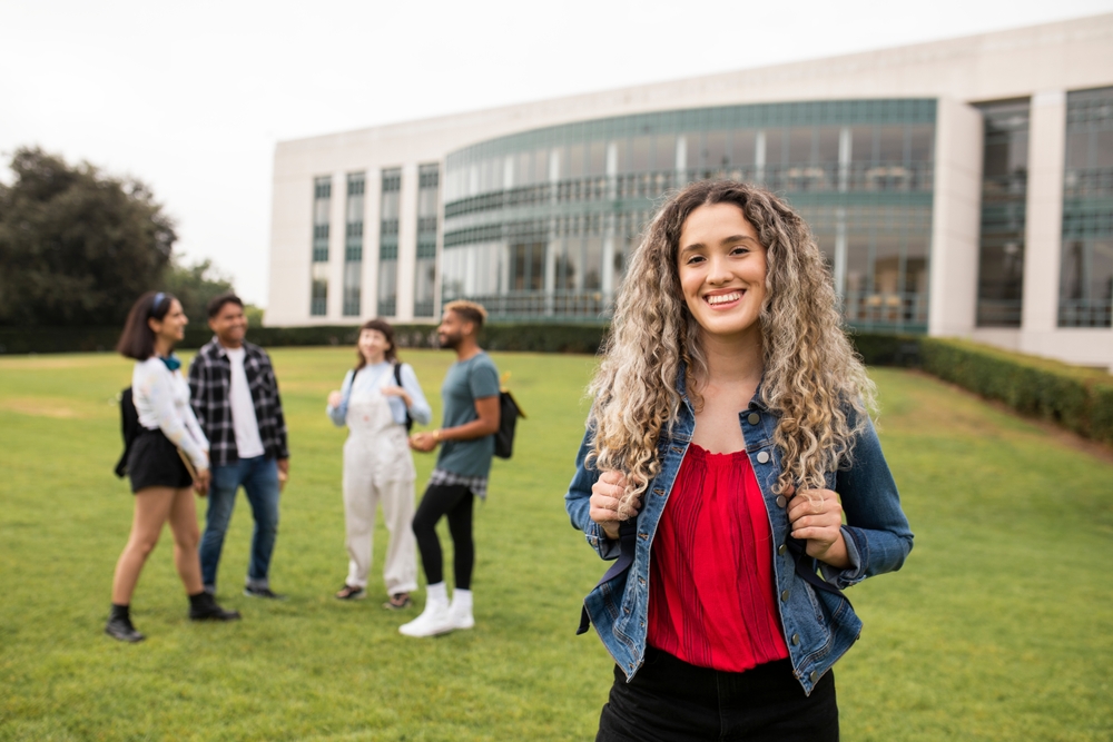 Jovem mulher segurando a alça da mochila e sorrindo para a câmera, com quatro estudantes conversando em grupo ao fundo, em um gramado de campus moderno.