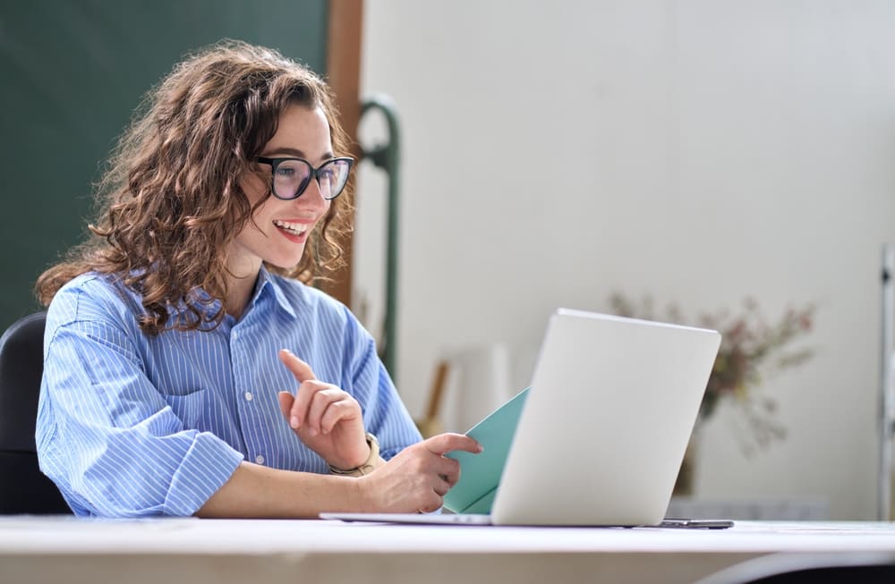 Mulher sorrindo em frente ao laptop, segurando um caderno e em ambiente tranquilo fazendo aulas de inglês para obter sua proficiência no idioma.