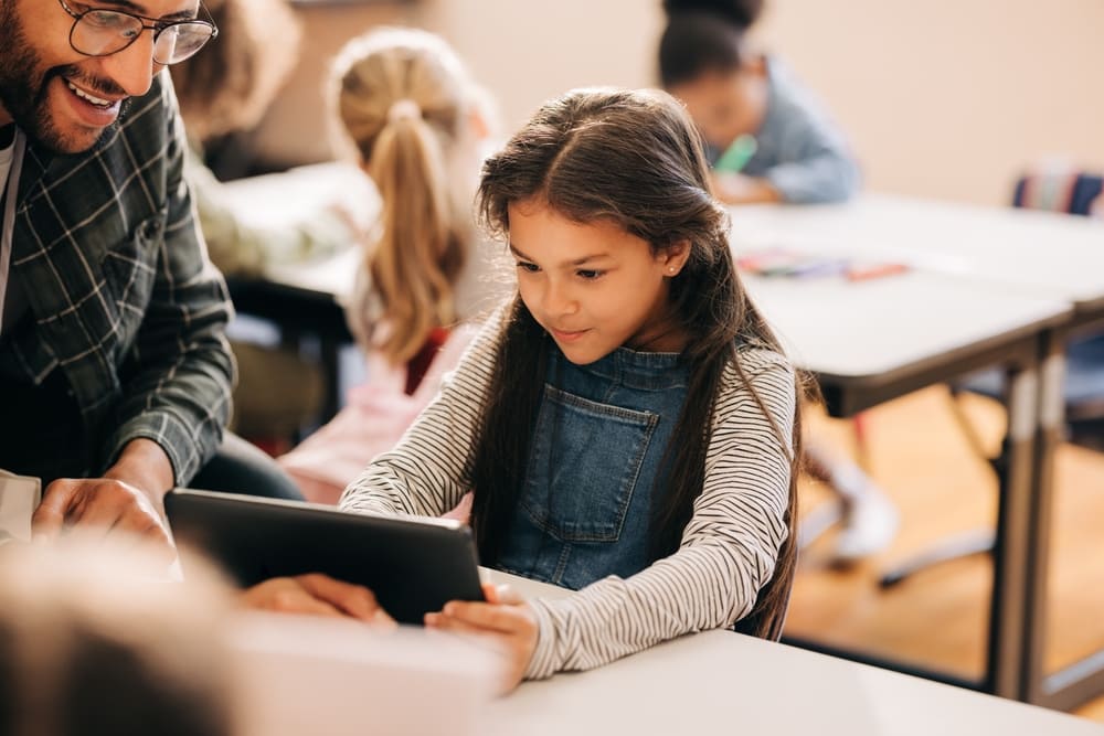 Criança aprendendo com tablet na sala de aula com apoio do professor, exemplo de letramento digital.