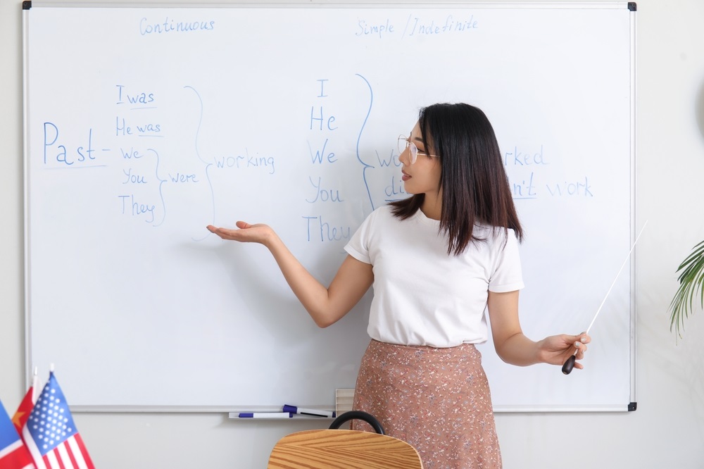 Professora ensina tempos verbais em inglês no quadro branco, ilustrando a importância da língua inglesa na comunicação global e na educação.