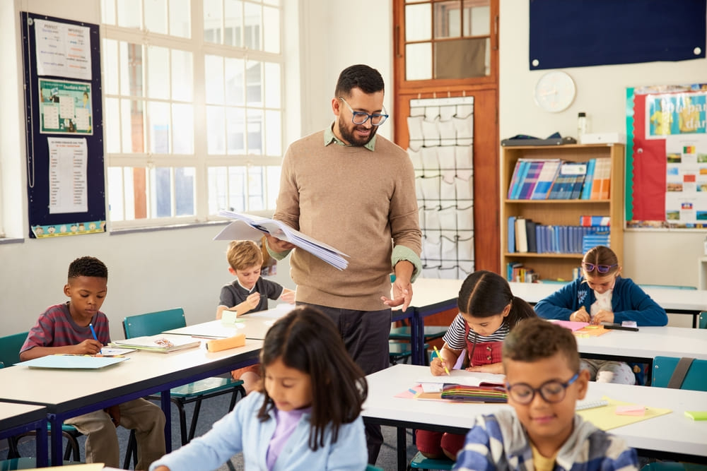 Professor acompanha alunos em atividades escritas em sala de aula, representando práticas relacionadas às tendências pedagógicas.