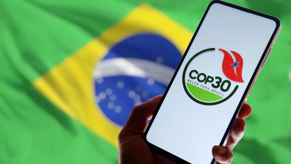 Logo da COP 30 no celular, com bandeira do Brasil ao fundo.
