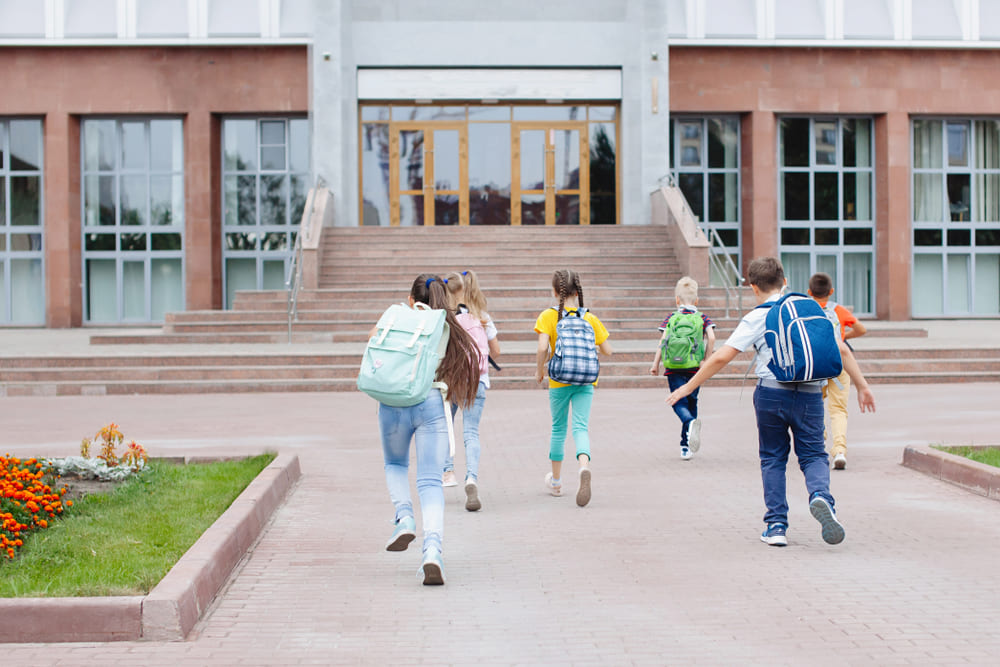 Infraestrutura da escola com acesso seguro: crianças chegam à entrada com escadas, corrimão e pátio bem cuidado.