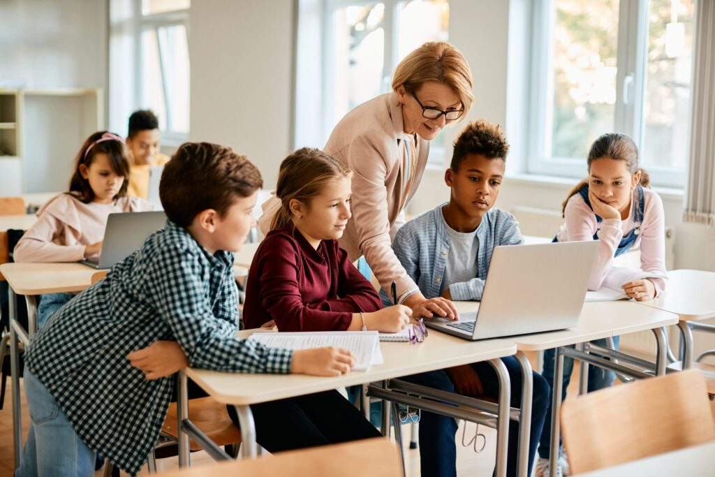 Crianças em sala de aula aprendendo com a professora em um laptop, exemplificando a inclusão digital na educação.