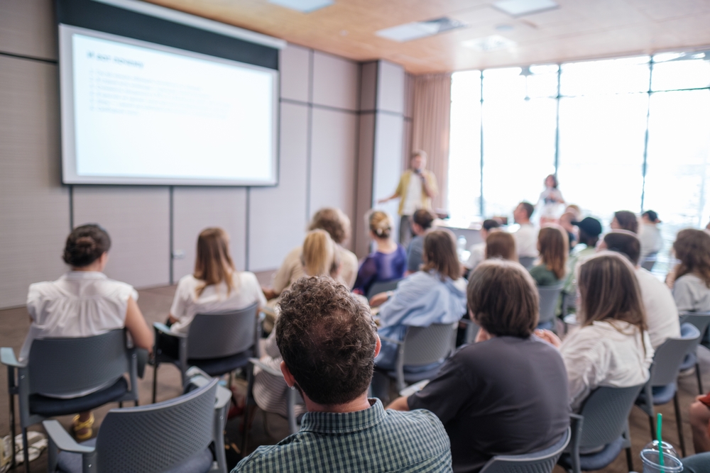 Pessoas assistindo a uma apresentação em sala de conferência, com foco em gestão pedagógica.