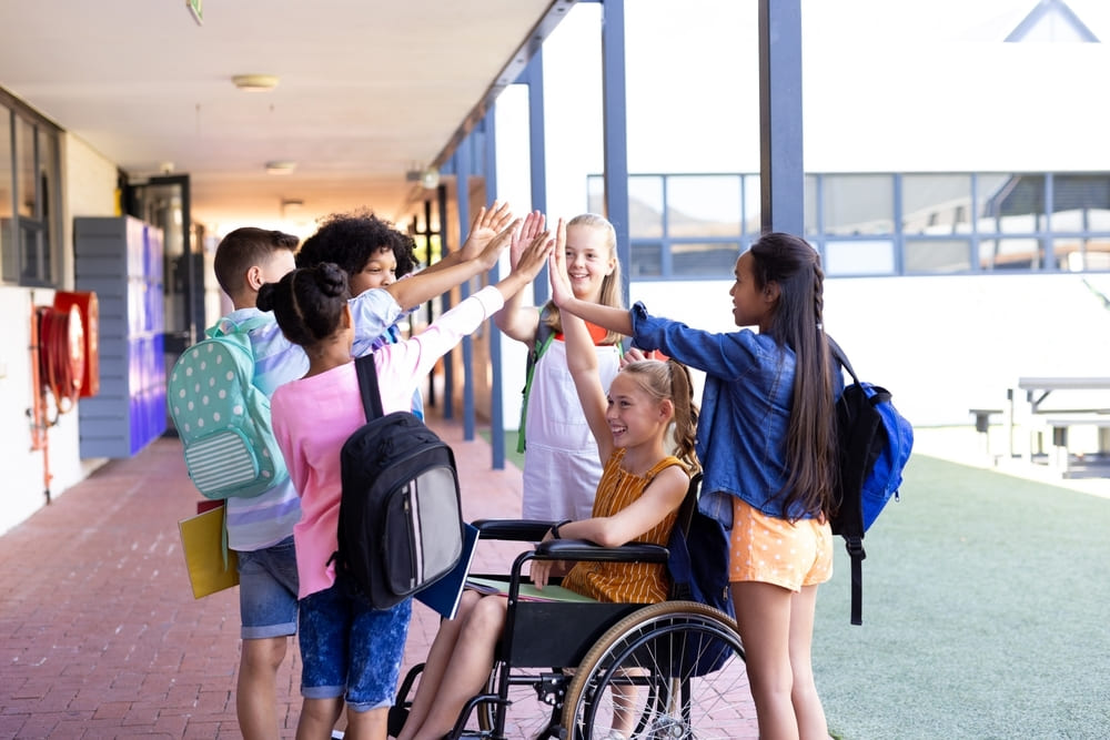 Crianças em corredor de escola celebrando juntas, incluindo uma aluna em cadeira de rodas, representando respeito às diferenças e inclusão escolar.