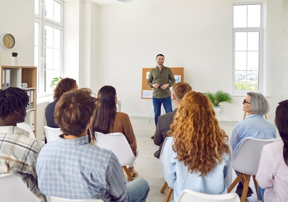 Dinâmica para reunião de pais com um professor falando para um grupo de responsáveis sentados em uma sala clara e organizada.