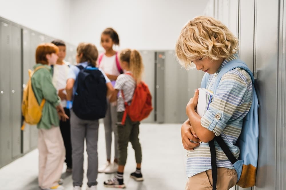 Menino isolado em corredor escolar enquanto colegas conversam em grupo, em cena que revela a importância de prevenir o bullying desde cedo na escola.