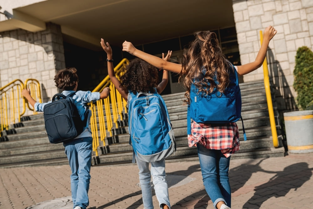 Crianças caminhando para a escola, com mochilas, comemorando o retorno às aulas.