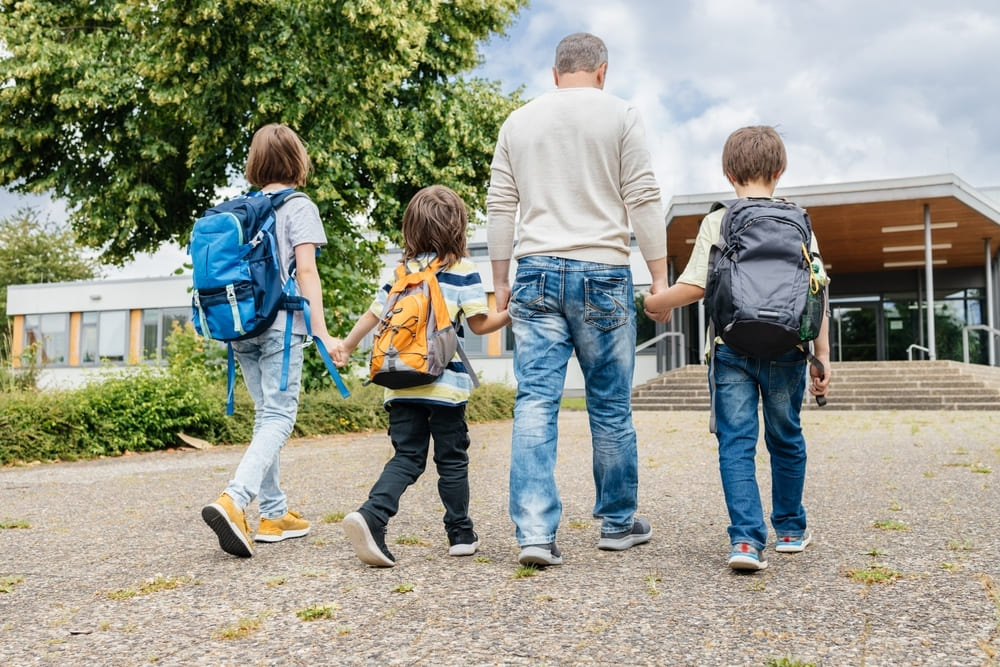 Pai e filhos caminhando de mãos dadas em direção à escola, representando a adaptação escolar.