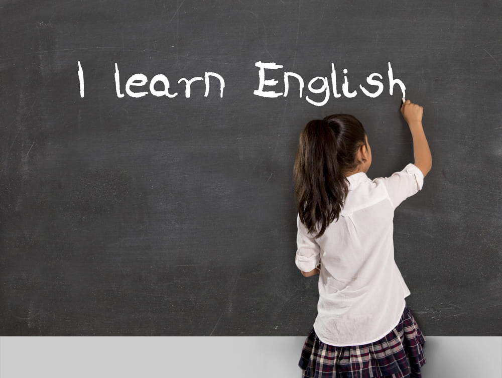 Criança escrevendo "I learn English" no quadro negro, representando o aprendizado de inglês em uma escola bilíngue.