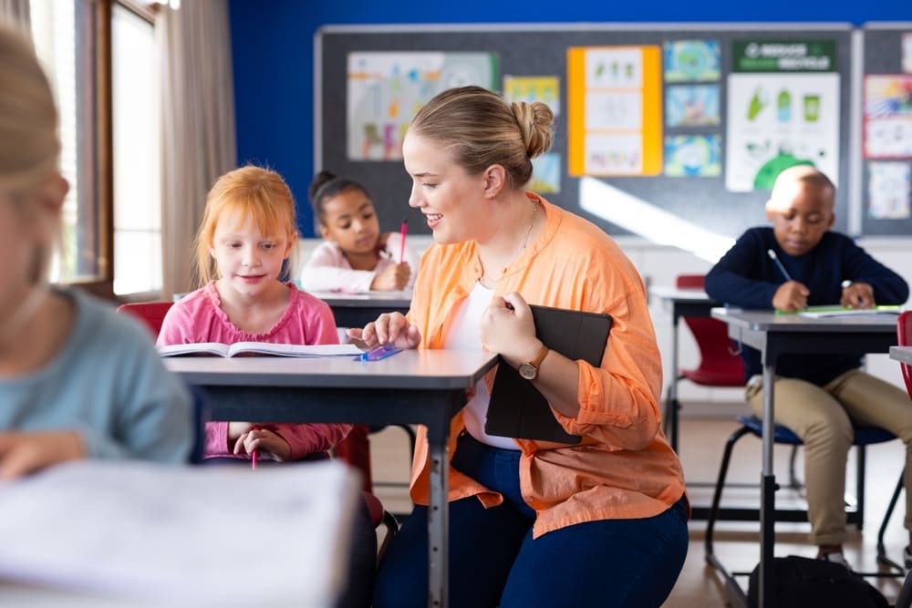 Professora interagindo com aluna em sala de aula, promovendo a comunicação escolar, enquanto outras crianças estão atentas às suas atividades.