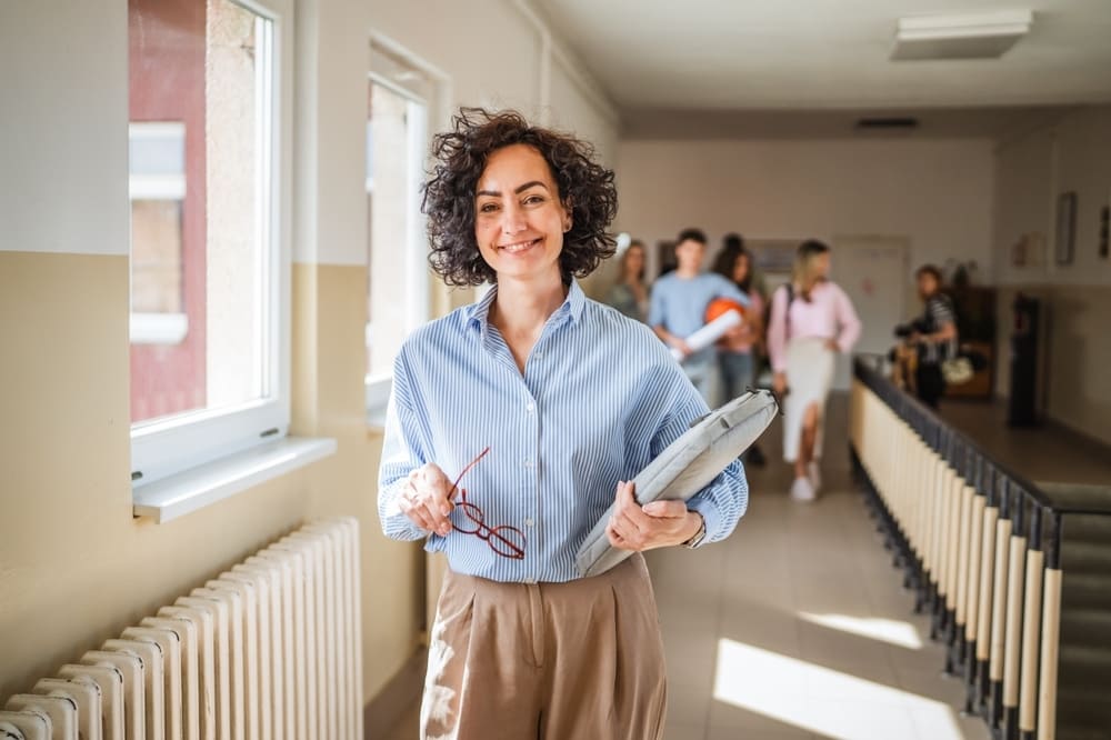 Professora sorridente com laptop e óculos em corredor escolar, com alunos ao fundo, representando um ambiente de aprendizado positivo e colaborativo.