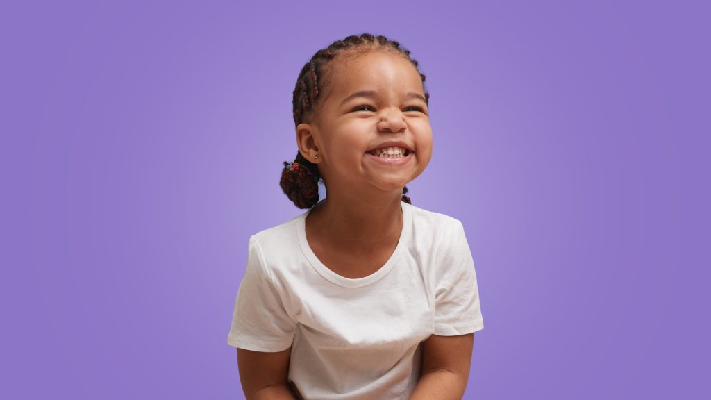 menina de 2 anos sorrindo como se olhasse para alguém. Ela tem a pele negra, cabelo trançado e veste camiseta branca em um fundo roxo.