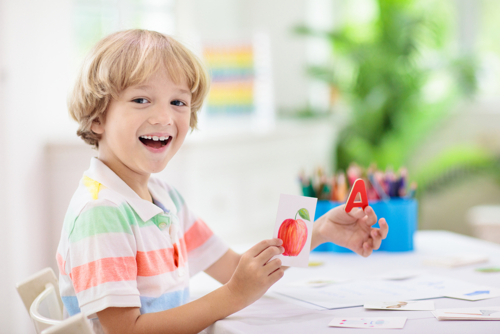 menino de 2 anos e loiro está sorrindo animado. Ele tem em uma mão um cartão com uma maçã e, na outra, um cartão com a letra A.