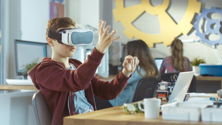 menino de 10 anos sentado na sala de aula usa óculos de realidade virtual e gesticula no ar