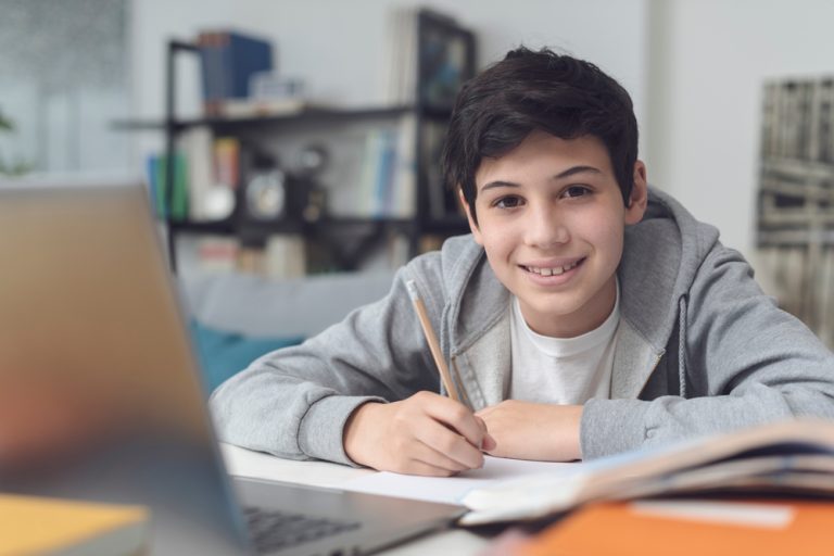 menino adolescente sentado, escrevendo em um caderno com um notebook à frente, sorrindo para a câmera