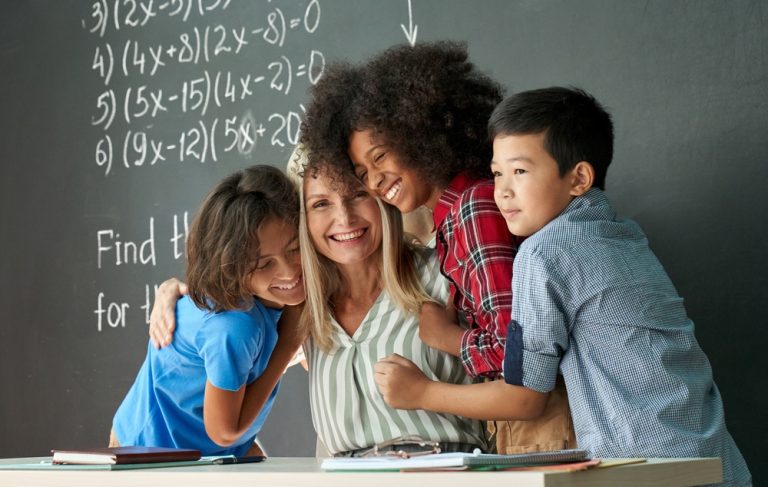 Professora abraçada com três alunos com cerca de 10 anos de idade em sua mesa, em frente à lousa de giz.
