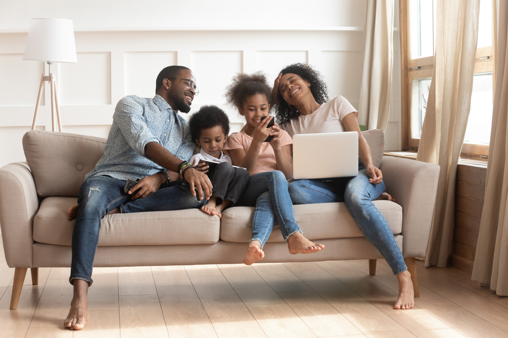 Família de pai, mãe e duas crianças sentados no sofá usando notebook, tablet e celular, todos sorrindo.