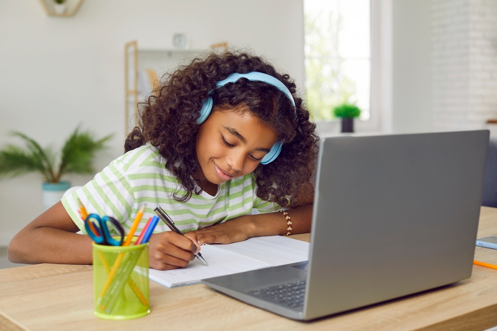 Menina de 10 anos estudando com caderno, notebook e fone de ouvido.