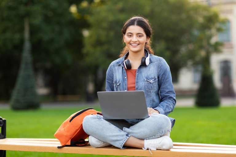 Estudante menina adolescente sentada ao ar livre sorri para a câmera com notebook no colo, headphones no pescoço e mochila ao lado.