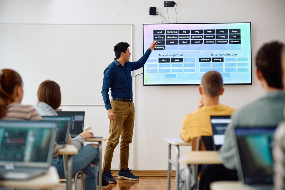 Professor apresenta conteúdo em sala de aula apontando para uma tela digital.