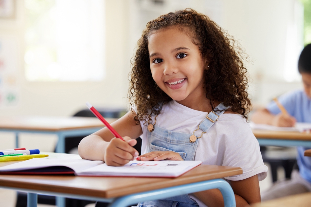 Menina de 6 anos escreve no caderno sentada na carteira da escola sorrindo para a câmera