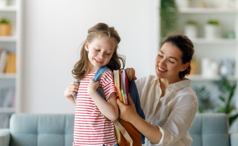 mãe ajuda filha pequena a colocar os materiais na mochila para ir à escola.