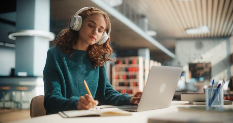 garota adolescente com cabelos ondulados e castanhos na altura dos ombros, veste moletom fechado verde escuro. Sentada na mesa de uma biblioteca, usa fones de ouvido brancos e escreve em um caderno enquanto usa o notebook.