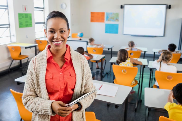 Professora negra de pele clara, cabelo preso, veste uma camisa salmão e cardigan bege claro. Ela sorri para a câmera, de pé, no meio da sala de aula, segurando um tablet nos braços. Ao fundo, crianças estão sentadas de costas nas carteiras.