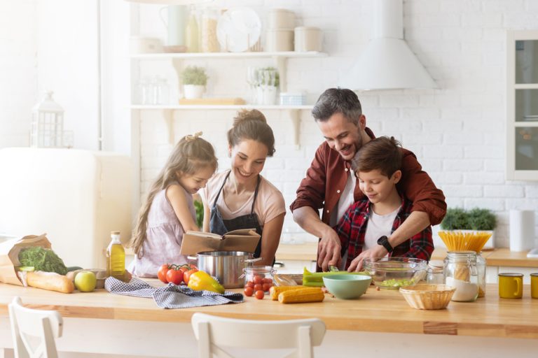 família de pai, mãe e duas crianças pequenas cozinhando juntos em uma bancada. Todos têm a pele branca, cabelo castanho e vestem roupas casuais.