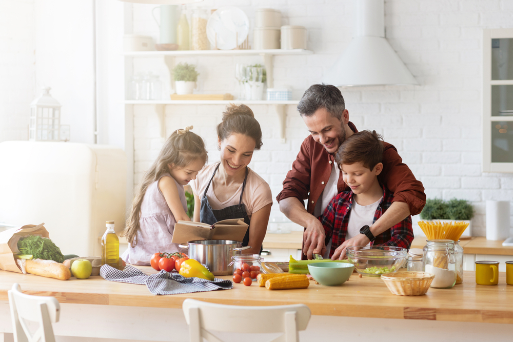 família de pai, mãe e duas crianças pequenas cozinhando juntos em uma bancada. Todos têm a pele branca, cabelo castanho e vestem roupas casuais.