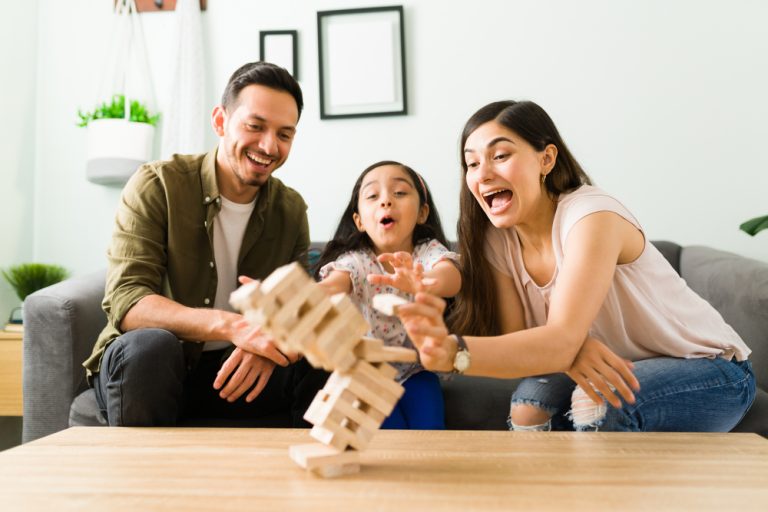 Pai, filha pequena e Mãe estão sentados no sofá e reagem com espanto e alegria ao ver que a torre de blocos de madeira que estão montando está começando a cair.