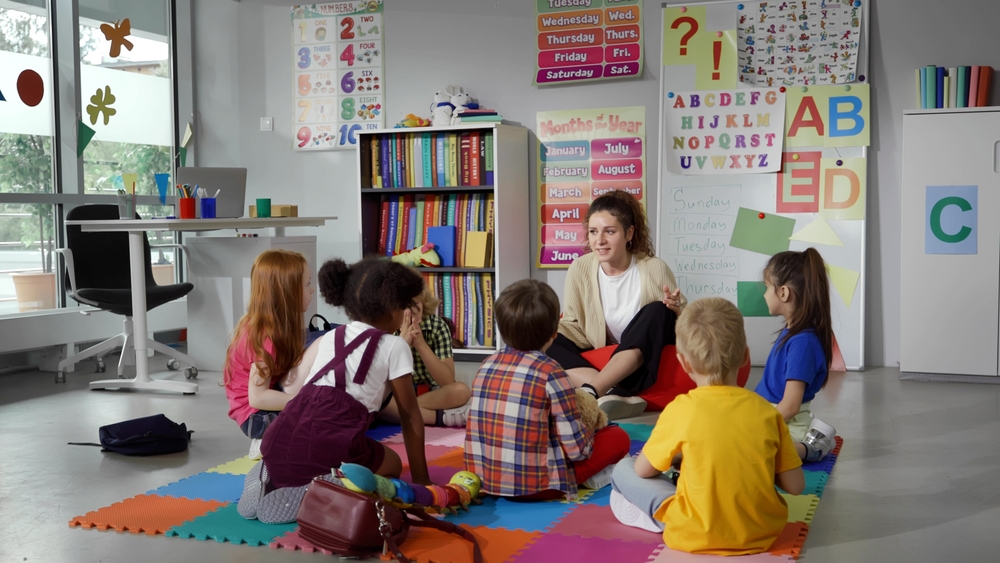 Crianças da Educação Infantil sentam em roda, no chão, junto com a professora.