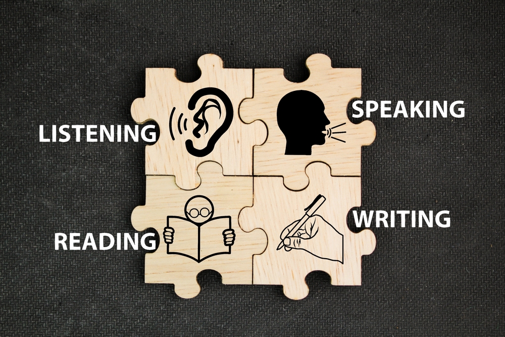 A imagem apresenta um conjunto de peças de madeira de quebra-cabeça com ícones e palavras que representam as quatro principais habilidades em inglês: listening (escuta), speaking (fala), reading (leitura) e writing (escrita). A composição mostra como essas habilidades se complementam no processo de aprendizagem da língua inglesa.