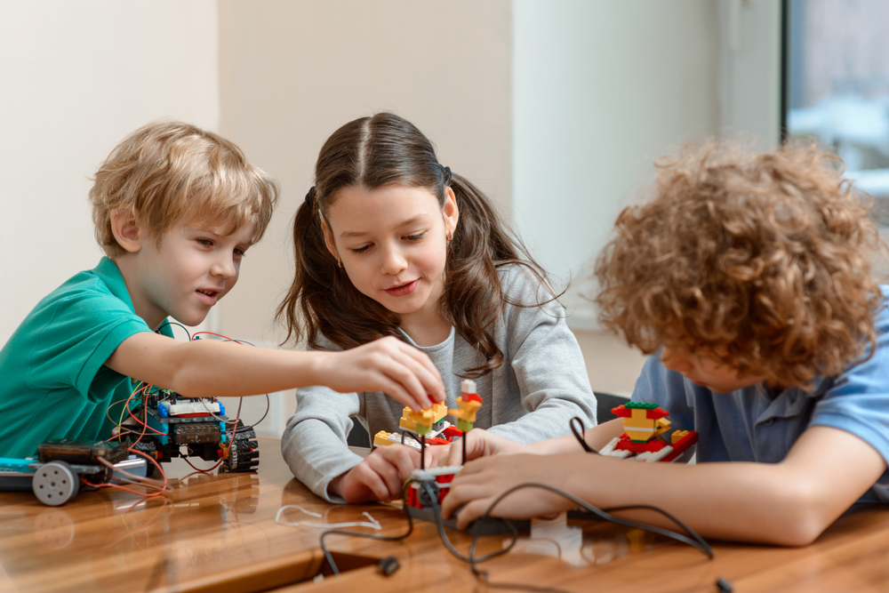 Dois meninos e uma menina, todos com cerca de 8 anos, brincam de montar um lego robotizado em cima de uma mesa de madeira.
