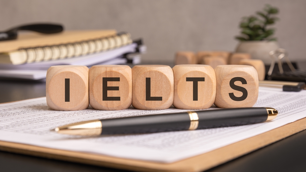 Blocos de madeira com as letras IELTS sobre uma mesa com documentos, caneta e caderno.