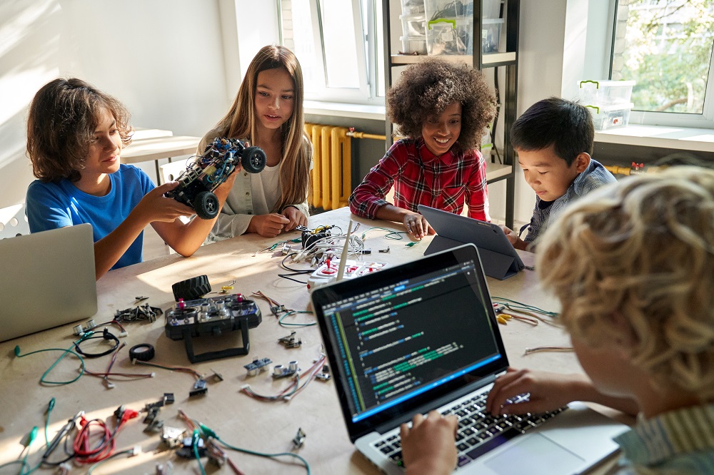 Crianças em atividade prática de programação e robótica, usando laptop, tablet e peças eletrônicas, representando o conceito de “pensamento computacional” no ensino fundamental.