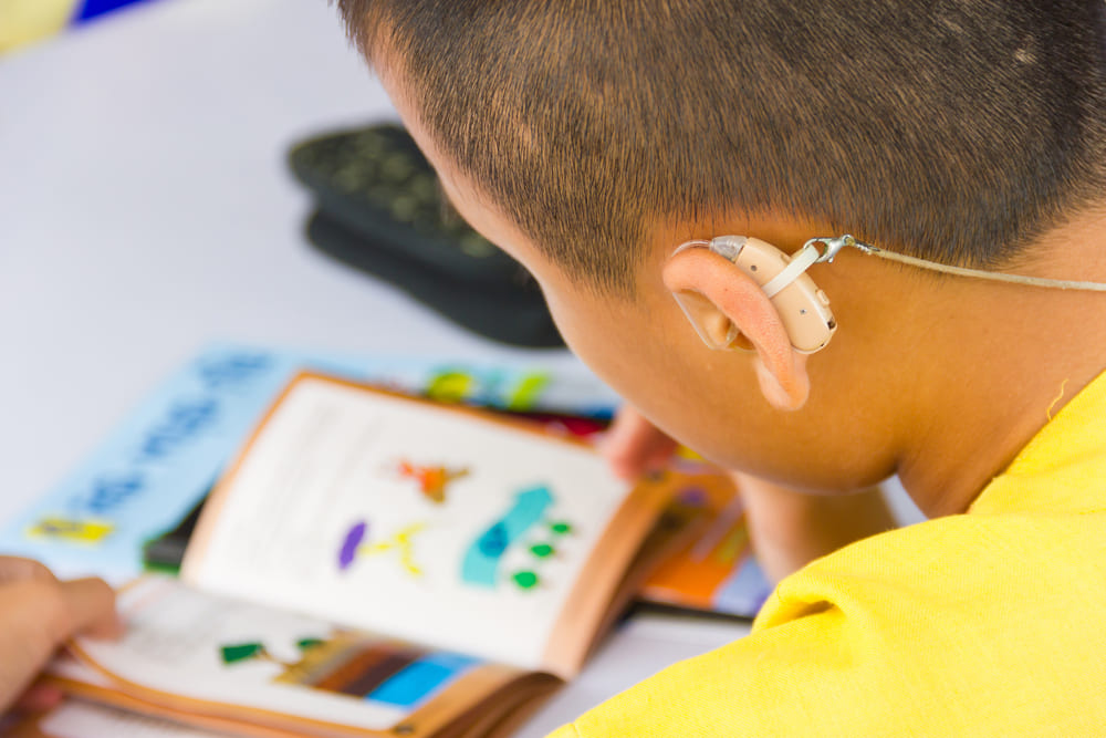 Criança com aparelho auditivo lendo um livro, ilustrando o uso de tecnologia assistiva na educação.