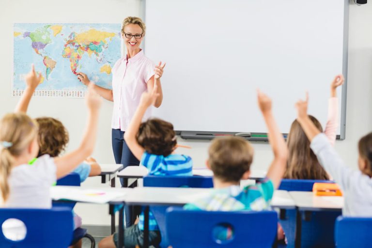 Professora aponta mapa; alunos levantam a mão em sala de aula, exemplificando uma abordagem pedagógica com participação.